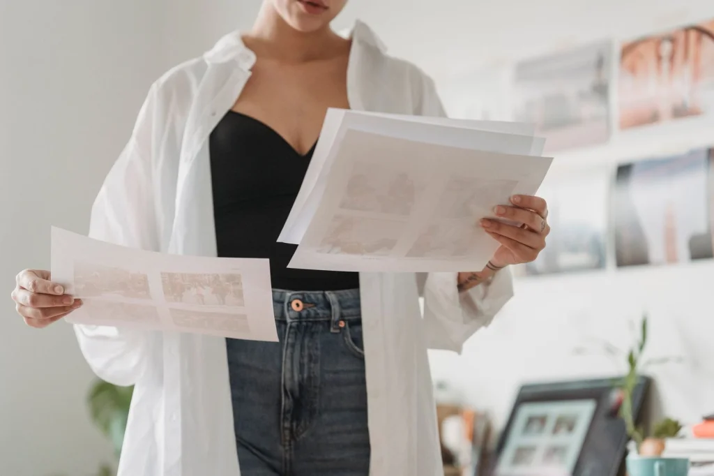 Interior designer in his 50s reviewing architectural photos on an iPad Pro, large format prints spread on drafting table, natural light from floor-to-ceiling windows, wide shot of modern office, editorial photography