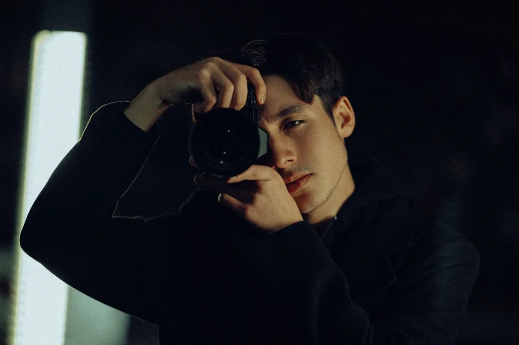 Professional photographer in his 30s with short dark hair testing AI background removal tools on multiple monitors showing before and after product photos, modern home office setup with camera equipment visible, warm desk lamp lighting from above, three-quarter view medium shot, lifestyle photography, shallow depth of field