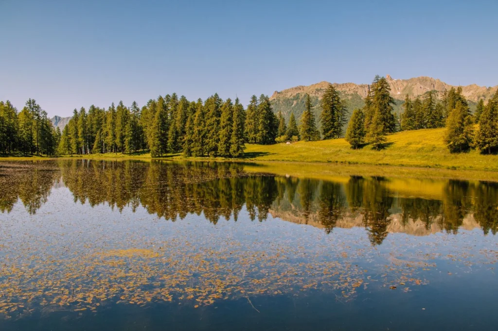 Mountain lake landscape with pine trees reflected in calm water, AI-expanded sky showing dramatic clouds and additional mountain peaks in the distance, golden hour lighting creating warm reflections, wide-angle landscape shot, cinematic nature photography with rich saturation