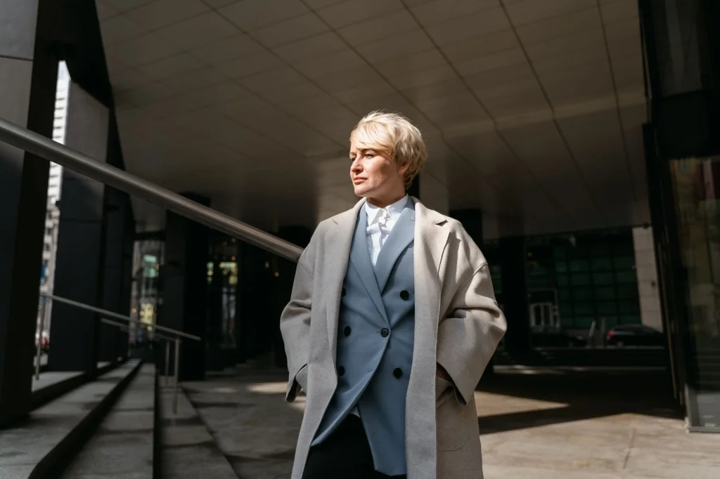 Professional woman in her early 30s with short blonde hair, wearing navy blue blazer and white blouse, slight confident smile, sitting at modern office desk with laptop, soft natural light from window, eye-level medium shot, corporate headshot style, shallow depth of field
