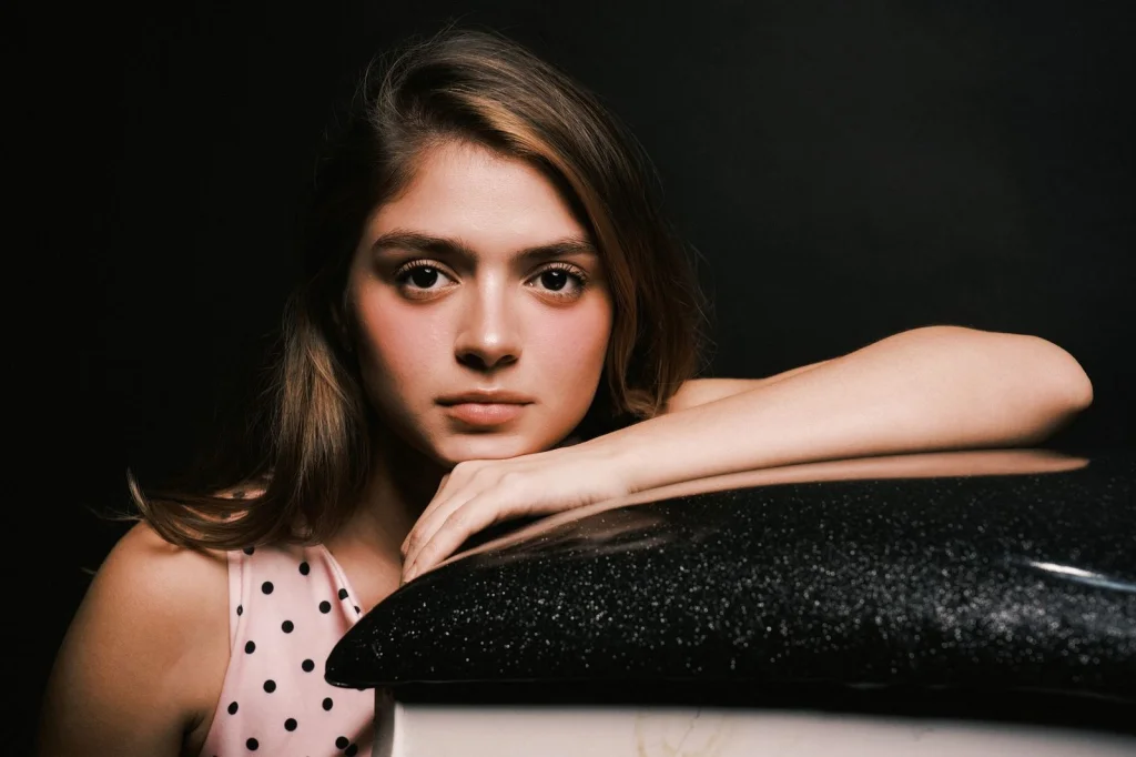 Woman in her late 20s with brown shoulder-length hair, sitting at a white vanity mirror, touching her hair thoughtfully, natural window light from the right, eye-level medium shot, lifestyle photography, soft focus background