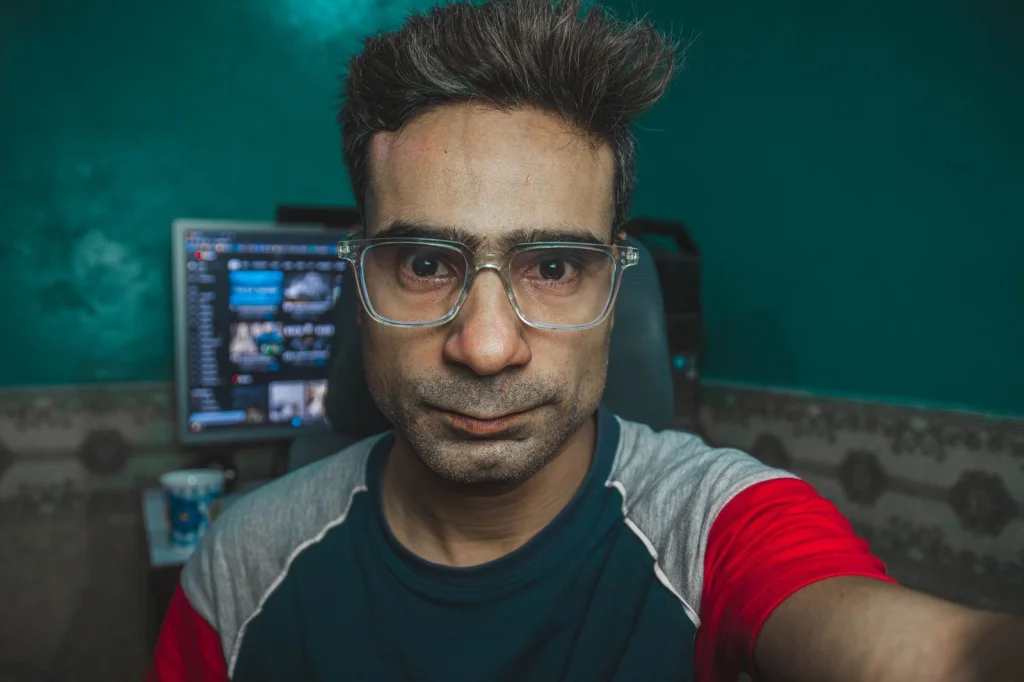 Computer setup showing a man in his 30s with glasses and a button-down shirt sitting at a desk with dual monitors displaying code, AI video generation interface visible on screen, cool LED lighting in background, medium shot from side angle, tech workspace photography