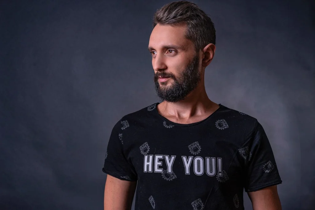 Man in his early 30s with short beard, dark hair, navy blue shirt, slight smile, white studio background, soft even lighting from front, chest-up portrait shot, professional headshot style