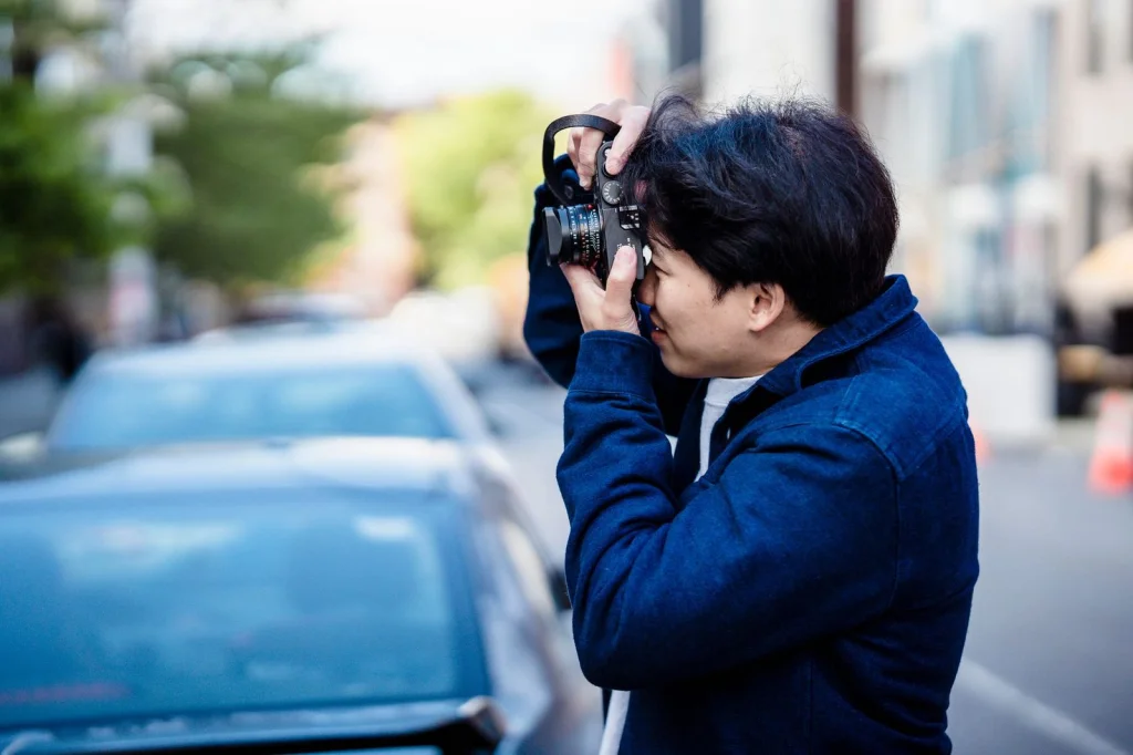 Professional photographer in his 40s with glasses, examining side-by-side photos on dual monitors showing AI-generated vs real portraits, modern home office with brick wall background, warm desk lamp lighting, over-shoulder medium shot, documentary photography, shallow depth of field
