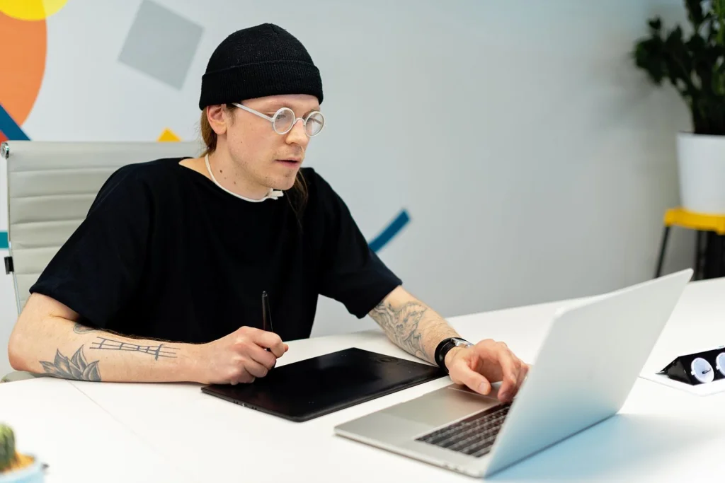 Digital artist in his 30s with glasses working on anime-style illustration on a drawing tablet, multiple reference images on wall, warm desk lamp lighting, over-shoulder view, creative workspace photography