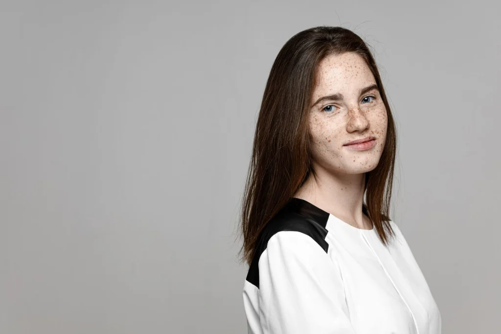 Woman in her late 20s with straight brown hair, wearing a white collared shirt, neutral expression looking directly at camera, sitting against plain white background, soft even lighting from front, head and shoulders portrait shot, passport photo style, sharp focus