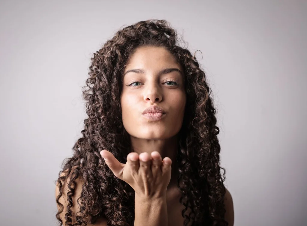 Woman in her 20s with natural 3B curls, shoulder-length dark hair, gentle smile, standing against a white wall background, soft natural light from window, eye-level medium shot, lifestyle photography, shallow depth of field