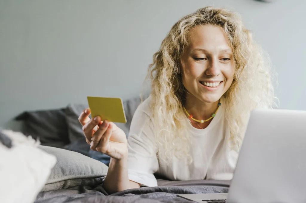 Young woman in her 20s with curly blonde hair wearing white sweater, sitting at laptop with Photoshop open showing transparent background result, bright modern kitchen with marble countertop, natural window light from right, medium shot at eye-level, lifestyle photography, soft focus background