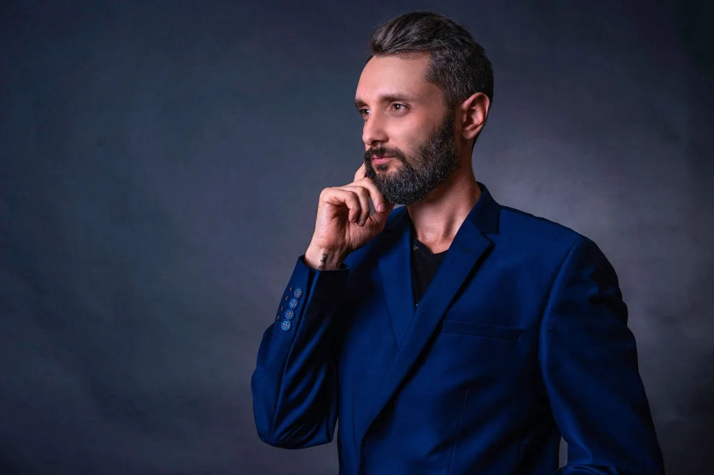Man in his 30s with beard, navy suit, confident expression, standing in a modern office with floor-to-ceiling windows, golden hour light streaming through glass, medium shot from chest up, editorial portrait, f/2.8 depth of field