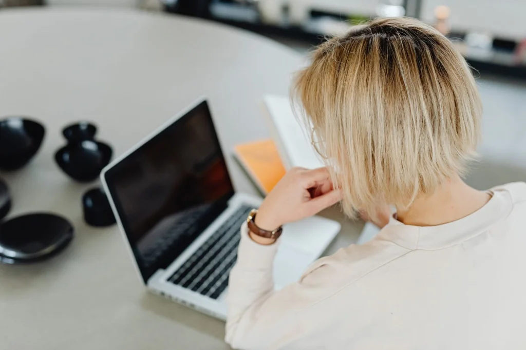 Woman in her 20s with blonde hair sitting at a desk, looking at a smartphone showing a face swap app interface, soft daylight from window, eye-level medium shot, editorial lifestyle photography, shallow depth of field