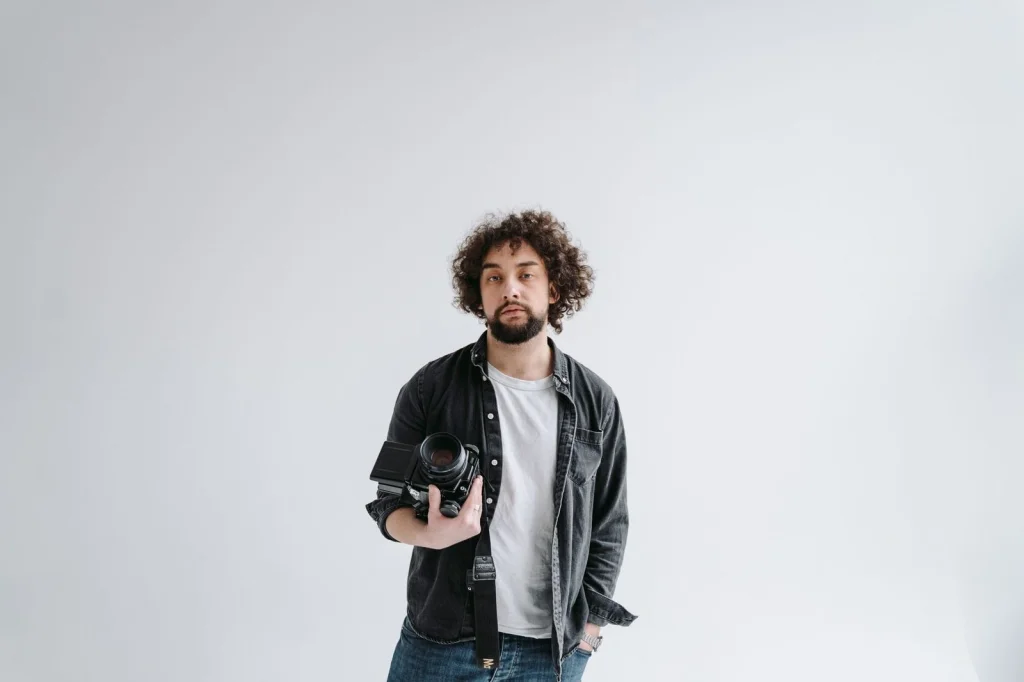 Professional photographer in his 30s with beard, black shirt, holding a DSLR camera, standing in a modern photo studio with lighting equipment, soft studio lighting, medium shot, editorial portrait