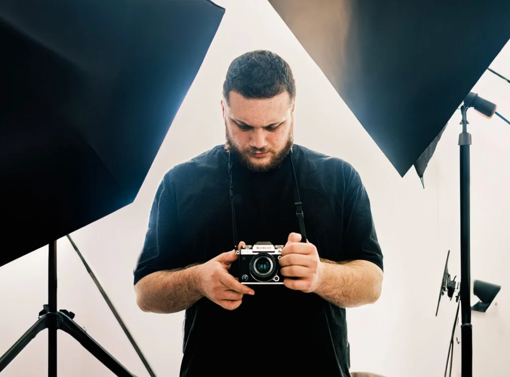 Professional photographer in his 40s with beard wearing black t-shirt, holding a DSLR camera, standing in a modern studio with photography equipment in background, professional studio lighting, waist-up shot, commercial photography
