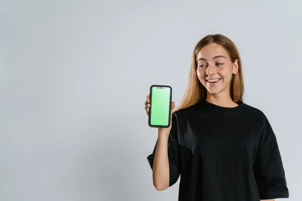 Young woman with straight brown hair holding a smartphone showing photo editing interface, sitting at a wooden coffee shop table with laptop open, soft natural lighting from large window, medium shot at eye level, lifestyle photography with warm tones
