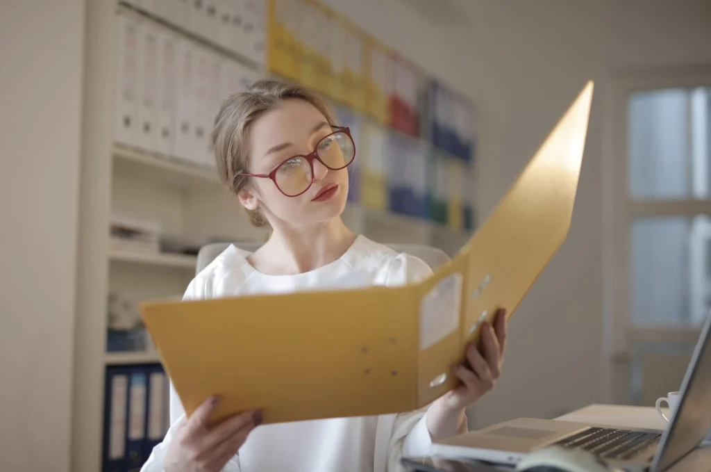 Creative professional woman in her thirties with glasses reviewing high-resolution printed photos spread across wooden table, natural window light, overhead flat-lay angle, editorial lifestyle, clean composition
