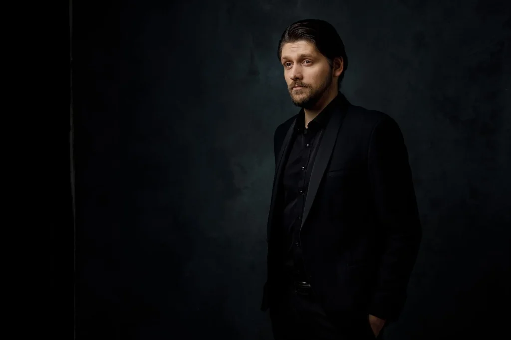 Professional businessman in his 30s with dark hair wearing navy blue suit and light blue dress shirt, confident expression, sitting at modern office desk with laptop, soft window light from the left, eye-level medium shot, corporate portrait photography, shallow depth of field