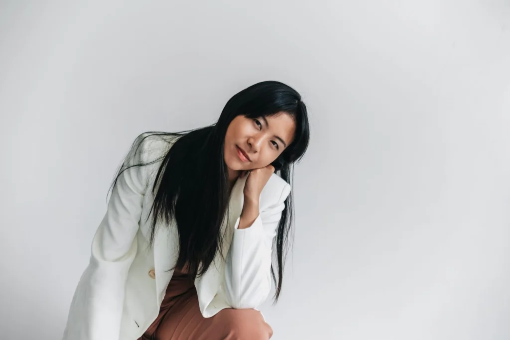 Woman in her 30s with black hair in a white blazer, confident smile, sitting at a modern office desk with plants visible, the AI-expanded background showing continuation of the office space with bookshelves and windows, soft natural lighting from the right, medium shot at eye level, professional corporate photography style