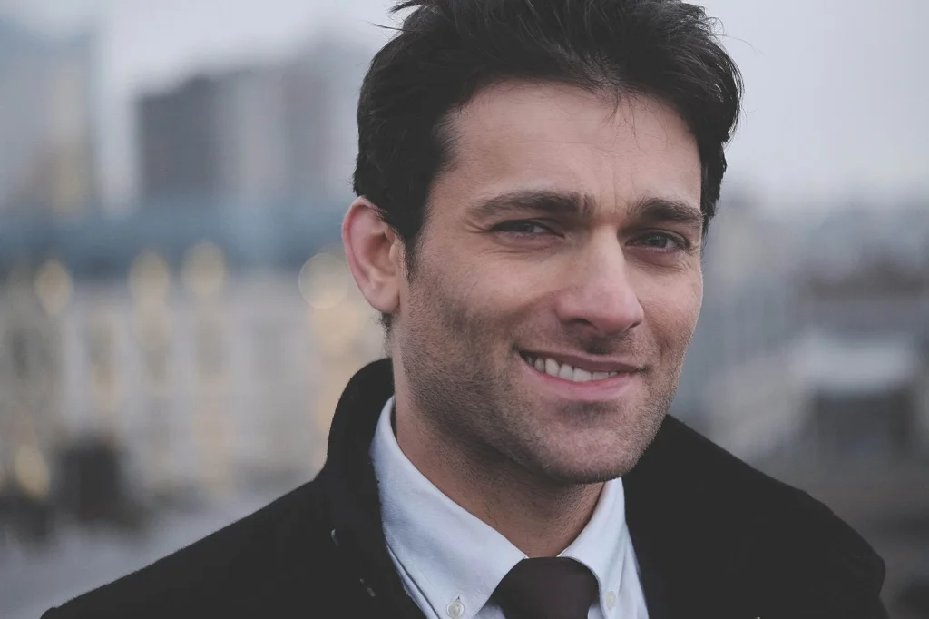 Professional man in his early 30s with dark hair and stubble, confident smile, wearing charcoal grey suit jacket with white dress shirt, hands clasped in front, modern office lobby with glass and steel elements, natural overhead lighting, waist-up shot at eye level, corporate lifestyle photography