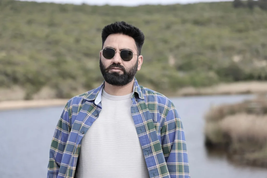 E-commerce product photographer in his 30s with beard, editing photos on a large monitor, removing backgrounds from white sneakers, modern studio with white backdrop and professional lighting, warm desk lamp lighting, over-shoulder view showing screen work, commercial photography aesthetic