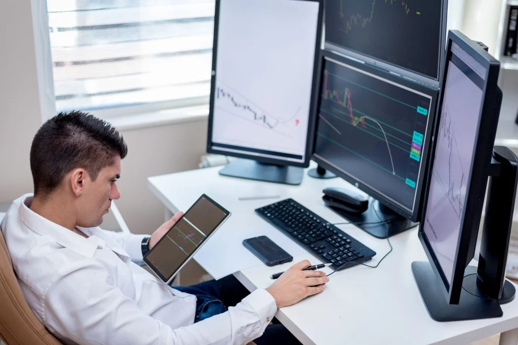 Professional photographer in her 40s sitting at a desk with multiple monitors showing before and after photos of background removal, organized workspace with camera equipment, natural window lighting, medium shot from the side, editorial workplace photography