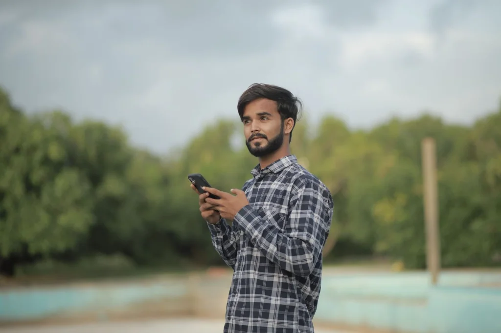 Young man in casual blue shirt holding smartphone, taking selfie with bright smile, modern office background with plants, natural window lighting, medium shot at eye level, lifestyle portrait photography
