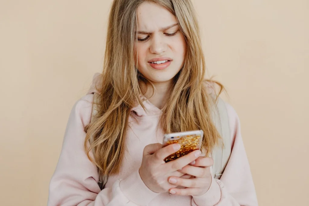 Young woman in her 20s with blonde hair holding a smartphone, smiling while using a photo editing app, sitting in a bright cafe with large windows, natural daylight from the left, close-up shot at eye level, lifestyle photography, soft focus background