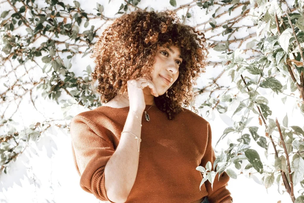 Woman in her early 30s with 3A curls in a burgundy sweater, testing a hair app on her phone, sitting at a cafe table with laptop, warm overhead lighting, three-quarter view close-up, editorial portrait, soft focus background
