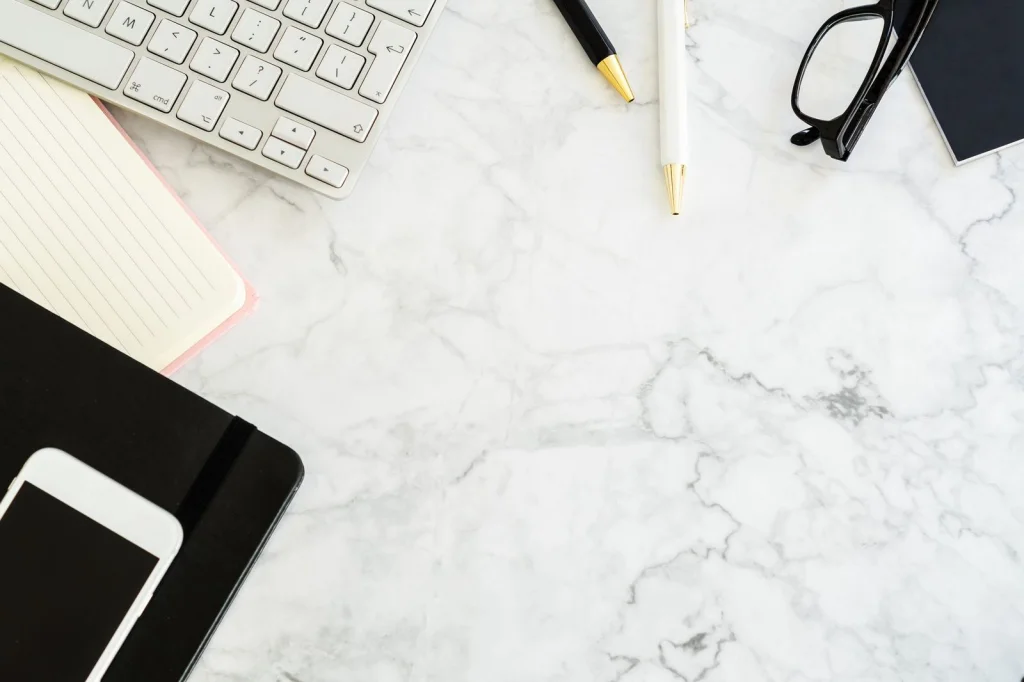 Flat-lay of a marble desk surface showing an Android smartphone displaying the Canva app with a product photo being edited, surrounded by a laptop, notebook with sketches, and a succulent plant, bright overhead lighting, top-down view at f/8, modern workspace photography