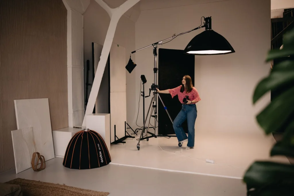 Professional photographer adjusting lighting setup for product photo shoot, camera on tripod, softbox lighting, white seamless backdrop, model holding reflector, studio environment, behind the scenes photography