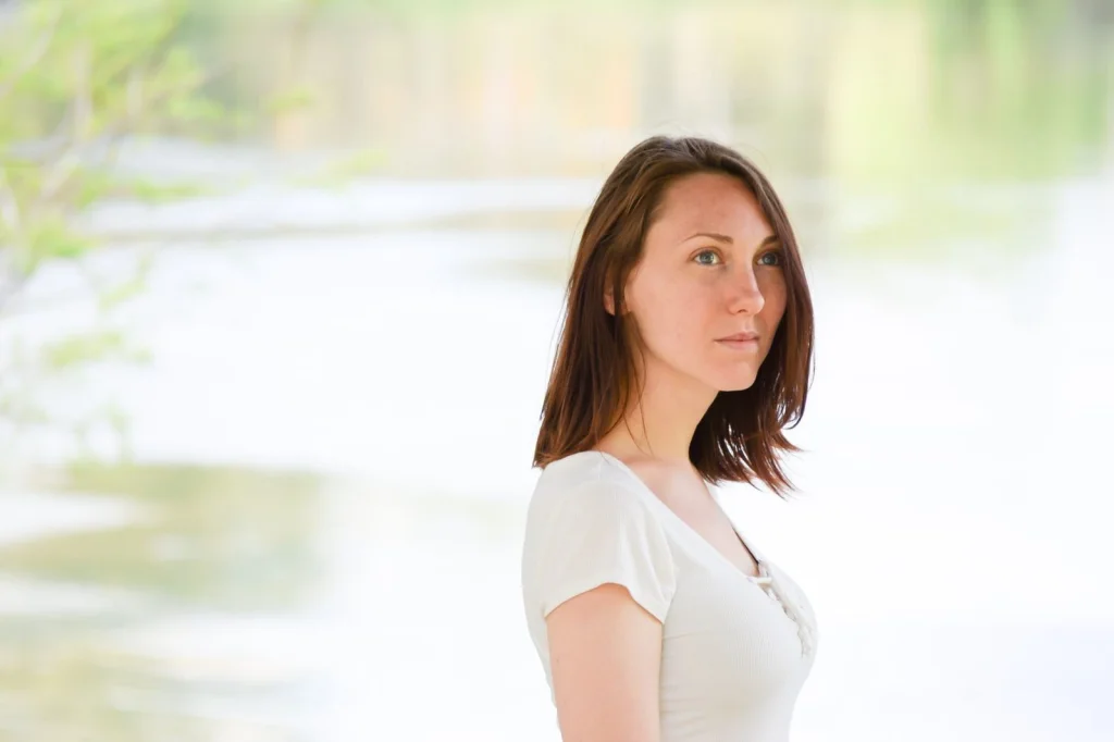 Young woman in her 20s with brown hair, white blouse, neutral expression, holding passport and printed photos, sitting at wooden desk with laptop, natural window light, medium shot, documentary style