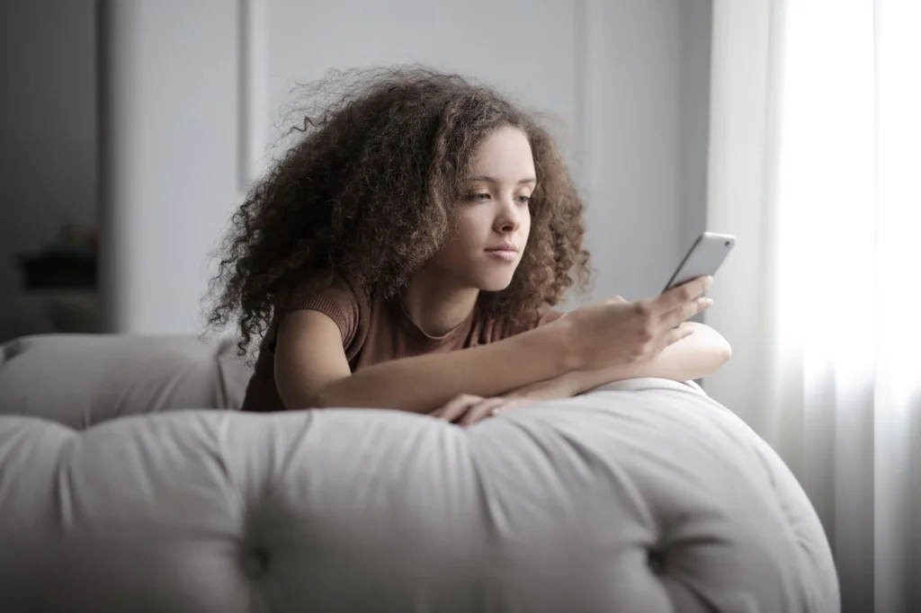 Teenage girl with curly brown hair, concentrated expression looking at phone screen, testing different hairstyles in a bright teen bedroom with fairy lights and colorful pillows, soft afternoon light from window, medium shot from slight above angle, lifestyle photography, warm tones