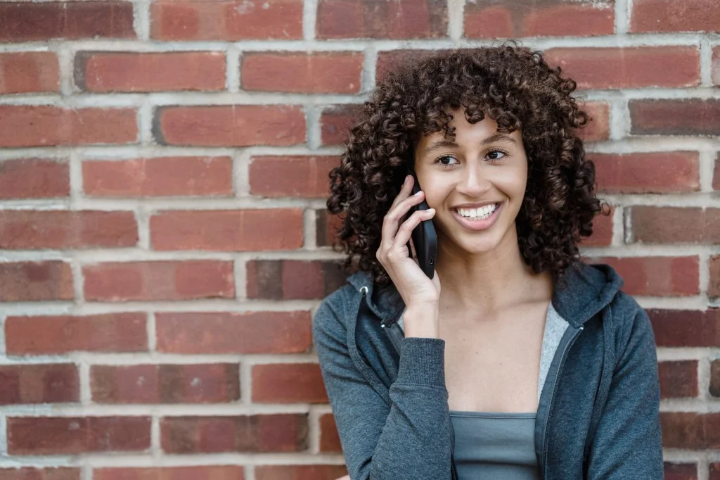 Young woman in her 20s with curly dark hair, wearing a white t-shirt, standing against a colorful painted wall background, holding a smartphone, soft natural lighting, medium shot at eye level, lifestyle photography