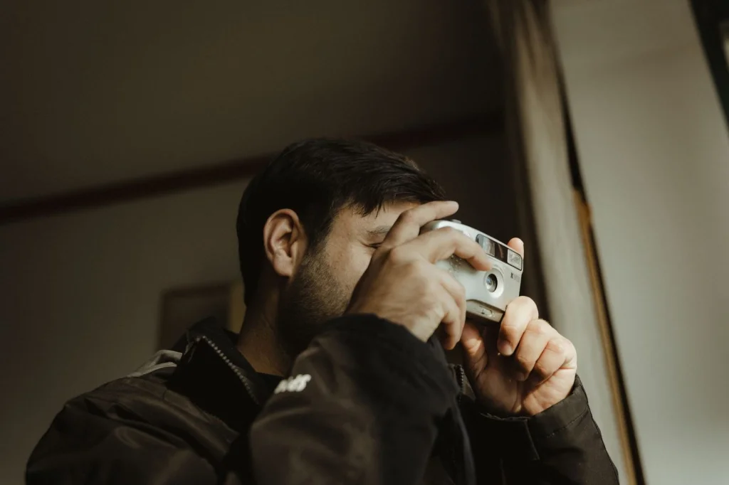 Professional photographer in his 30s with beard, testing AI image generation tools on a modern laptop, sitting in a bright studio with photography equipment in the background, natural daylight from large windows, eye-level medium shot, editorial workspace photography