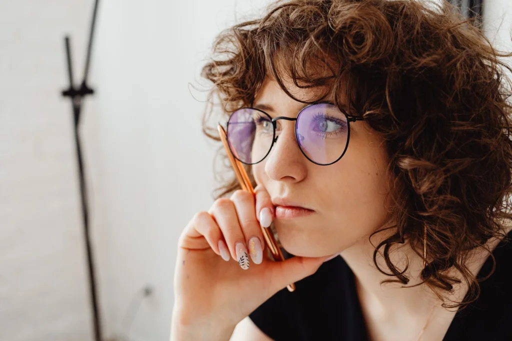 Woman in her 30s with curly brown hair, wearing a white blouse, standing in a busy cafe with blurred background elements, soft natural lighting from large windows, medium shot at eye level, lifestyle photography