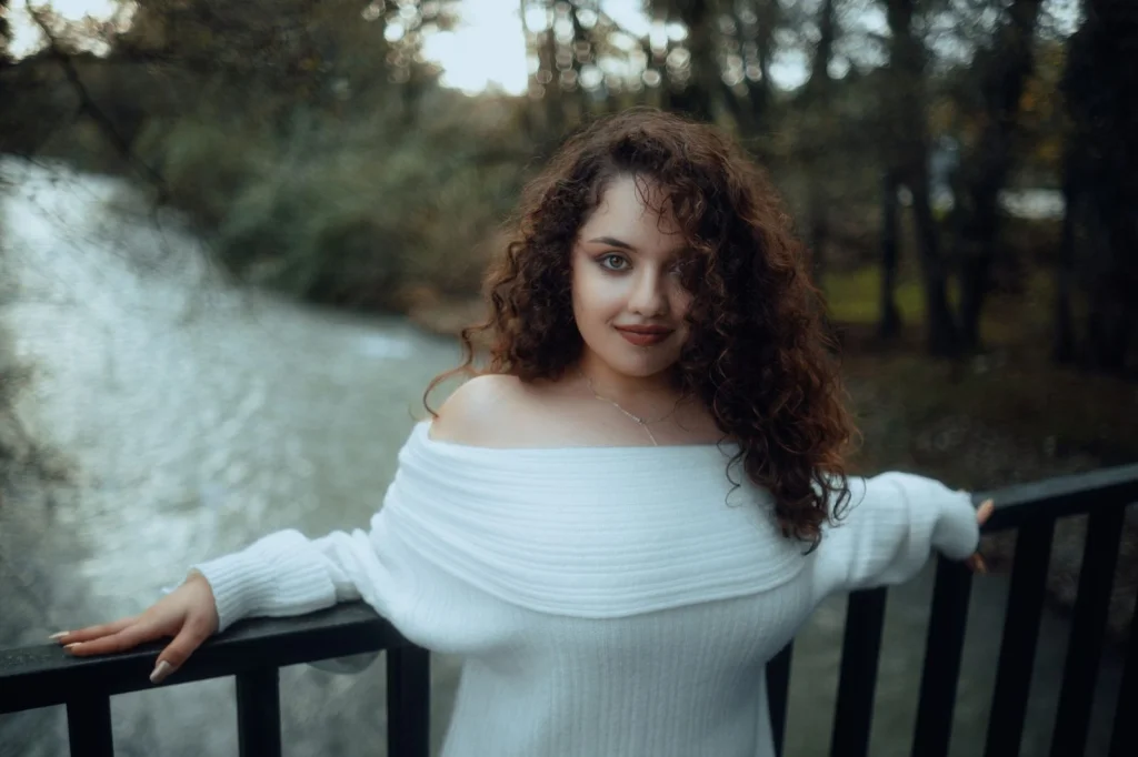 Young professional woman with curly brown hair, white blazer, gentle smile, standing against a complex busy office background with plants, natural daylight from large windows, medium shot at f/2.8, editorial portrait, shallow depth of field
