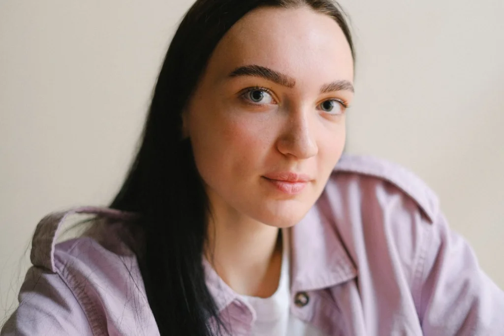 Woman in her late 20s with medium-length brown hair, neutral expression, sitting in front of a clean white background, soft diffused lighting, head and shoulders portrait shot, lifestyle photography, sharp focus