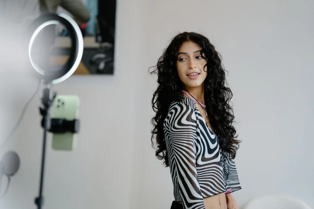 Young content creator in her early 20s with colorful hair and casual clothing editing video on laptop, multiple screens showing social media platforms, ring light and microphone visible, bright creative workspace with plants, over-shoulder shot, modern lifestyle photography