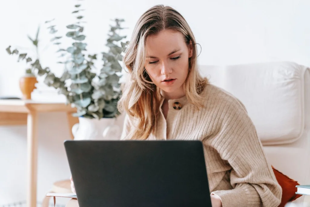 Graphic designer woman in her late 20s with blonde hair in a ponytail wearing a black hoodie, using Photoshop online on a large monitor showing background removal tool, modern office setup with plants, warm overhead lighting, over-shoulder view, professional workspace photography