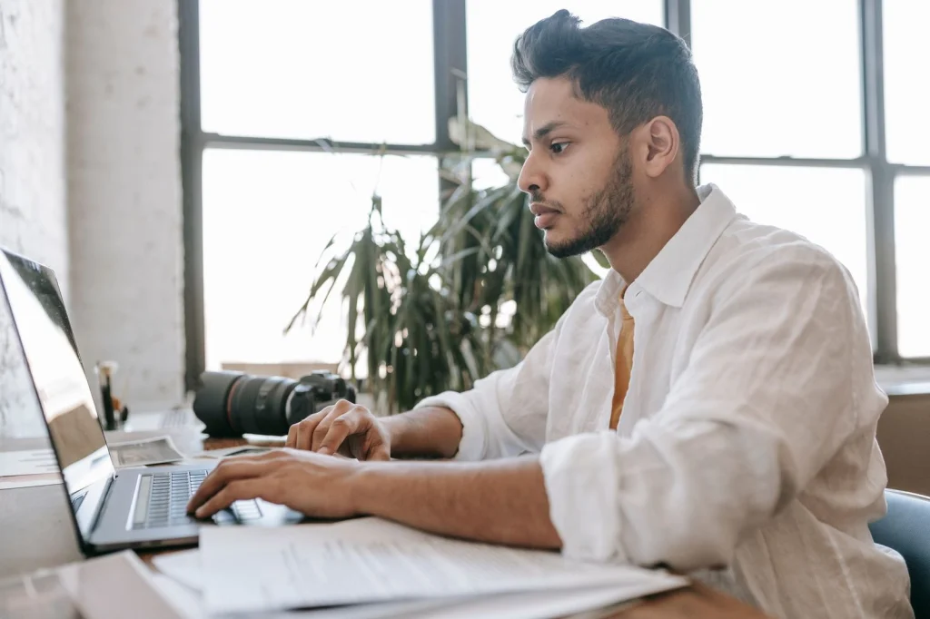 Professional photographer in his 30s with beard comparing two laptop screens side by side, one showing Photoshop interface and another showing an AI background removal tool, modern studio setting with camera equipment, warm LED lighting from above, overhead shot looking down, tech photography, sharp focus