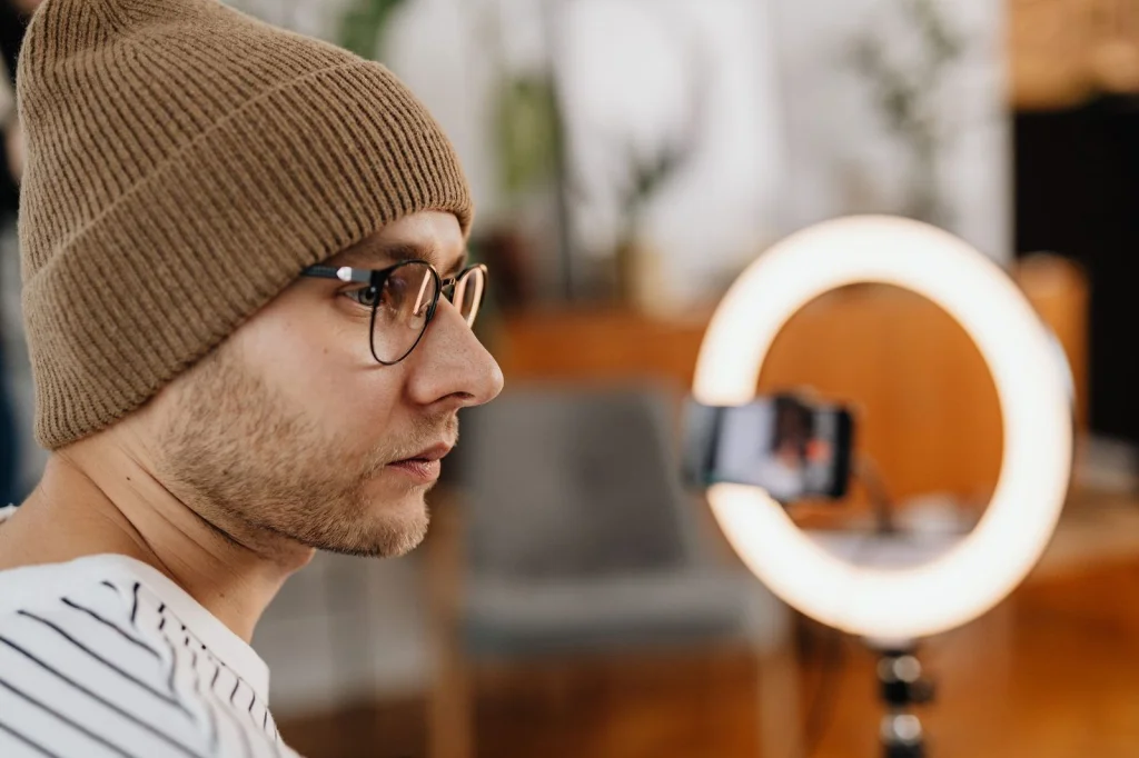 Social media manager in his 30s with glasses, creating visual content on dual monitors showing AI-generated images, modern office space with plants, natural lighting from window, side angle wide shot, contemporary workspace photography