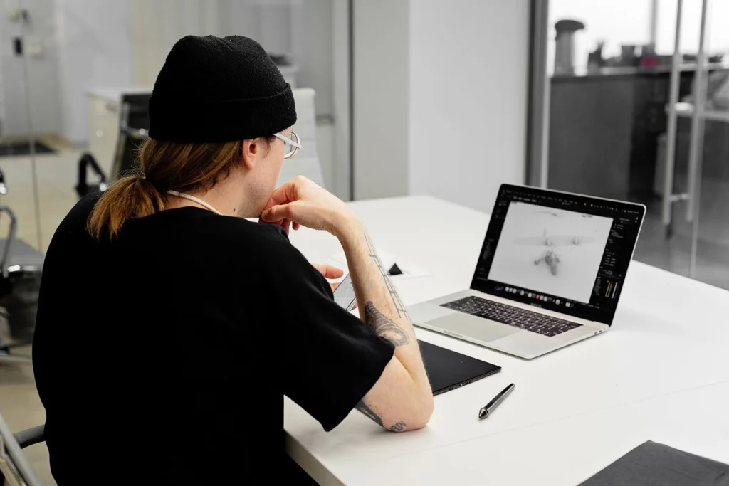 Freelance graphic designer in her late 20s with long black hair, wearing headphones and a gray hoodie, focused on dual monitors showing design software, working in a cozy home office with plants, warm ambient lighting, side angle medium shot, lifestyle photography
