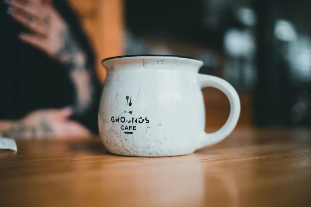 White ceramic coffee mug sitting on rustic wooden table with visible grain texture, AI-expanded background showing continuation of the wooden surface and warm kitchen environment with soft shadows, natural window lighting from above, overhead flat-lay perspective, lifestyle product photography with shallow depth of field