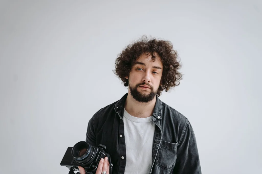 Professional photographer in his 30s with beard, wearing black shirt, holding DSLR camera, sitting at modern desk with multiple monitors showing photo editing software, warm studio lighting, medium shot, editorial style photography