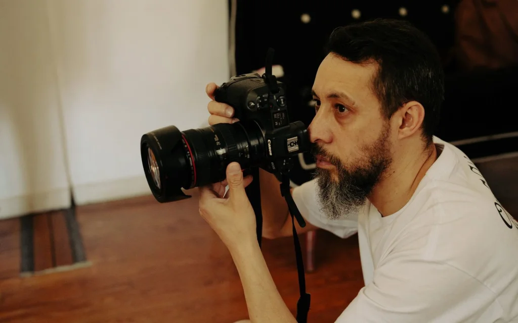 Professional photographer in his 30s with beard, analyzing before-and-after image quality on dual monitor setup showing blurry original and enhanced photo, modern home office with wooden desk, warm LED lighting from above, over-the-shoulder medium shot, editorial workspace photography