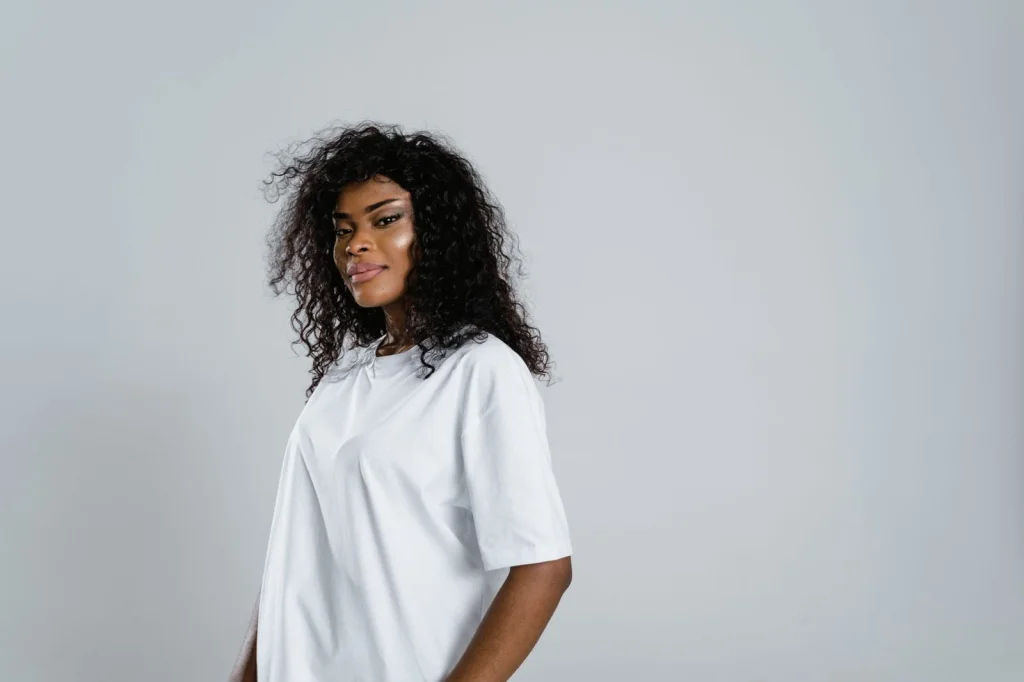 Woman in her 20s with curly hair wearing a white blouse, holding a smartphone at chest level, standing in a modern office with glass windows in the background, soft natural lighting from the side, waist-up shot at f/2.8, lifestyle photography