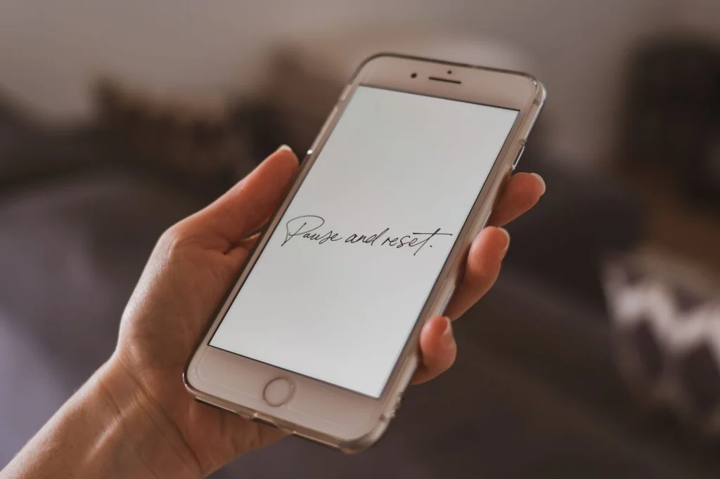 Close-up of hands holding a white iPhone displaying the Remove.bg app interface with a processed photo of a woman with background removed, sitting at a white desk with a coffee cup nearby, soft morning light through a window, overhead angle at f/4, clean product photography