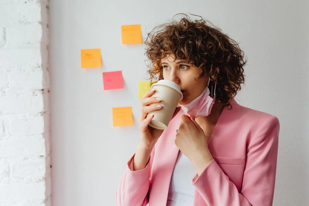Woman in her 20s with curly brown hair wearing a red sweater, arms crossed with confident smile, standing against a busy café background with blurred people, natural window light from the right, medium shot at chest level, lifestyle photography, shallow depth of field
