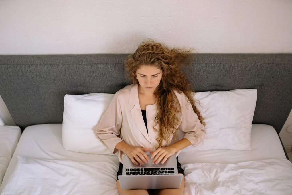 Freelance journalist in her 30s with curly hair, typing on laptop while analyzing news photos on second monitor, modern co-working space with plants, natural window light from right, medium shot from side angle, documentary lifestyle photography, natural color grading