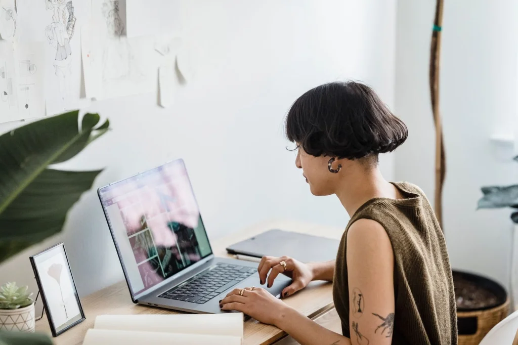 Young designer in his 20s with short brown hair, navy sweater, working on a laptop with design software open, sitting at a modern white desk, warm desk lamp lighting, over-shoulder view, workspace photography