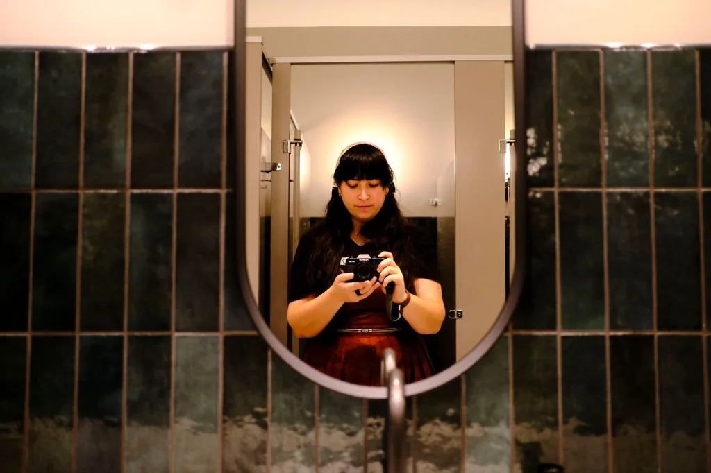 Young woman taking a selfie in her bathroom mirror, hair tied back in a loose ponytail, white t-shirt, natural makeup, bright bathroom lighting from vanity bulbs, close-up at f/2.8, lifestyle photography, crisp details