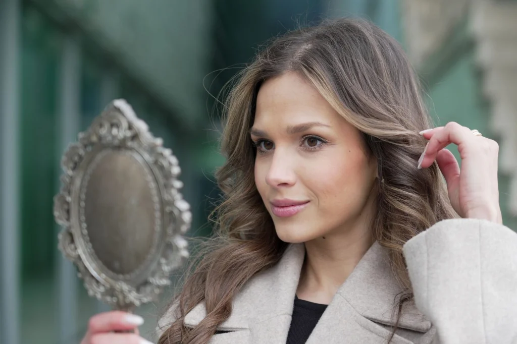 Woman in her 30s with brown hair comparing two photos side by side, before and after quality enhancement, sitting at a white desk with laptop and smartphone, soft natural light from window, eye-level medium shot, lifestyle photography